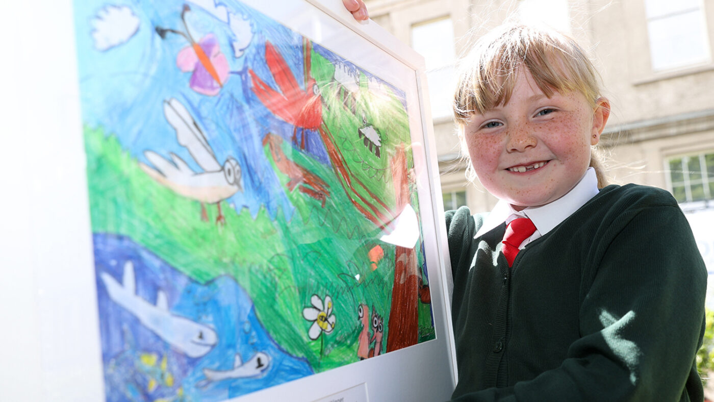 Ciara smiles while holding her framed drawing, which shows an environmental scene, with animals and insects in the sea, forest and sky.