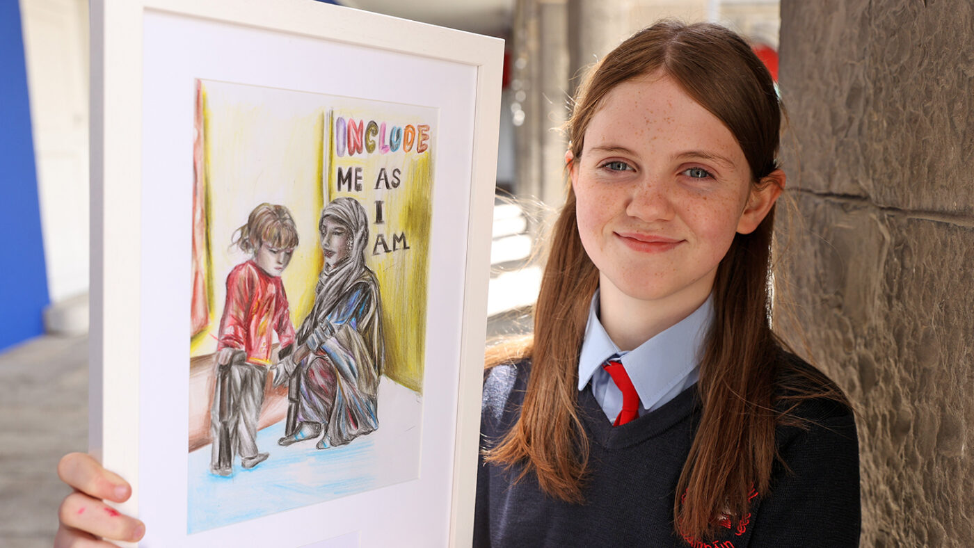 Aoife smiles as she holds her award-winning drawing, which shows a woman in a head scarf and a child. The text on the drawing reads 'Include me as I am'.