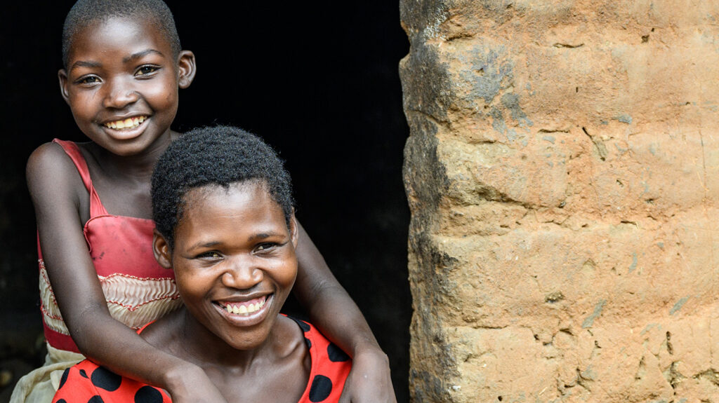 Mary hugs her mother in the doorway of their home. They're both smiling broadly.