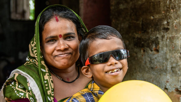 Sanjit smiles with his mother. He's wearing dark glasses. She's wearing a dark green headscarf.