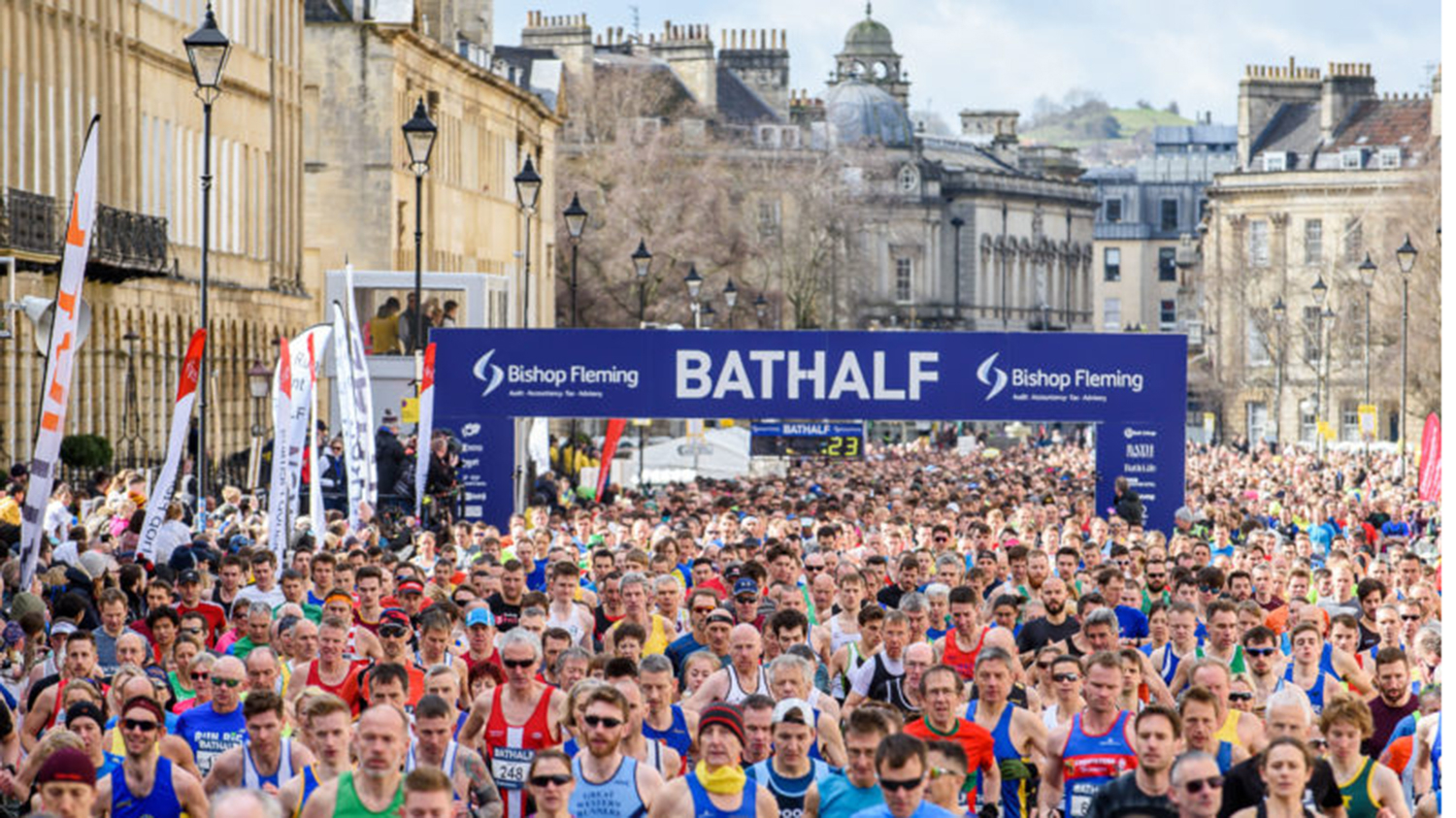 Hundreds of runners assembling under a large banner saying 'Bathalf'.