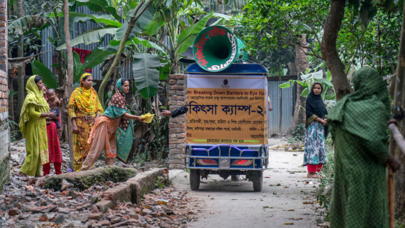 The tuk-tuk travels through a village, stopping to hand out flyers to women.