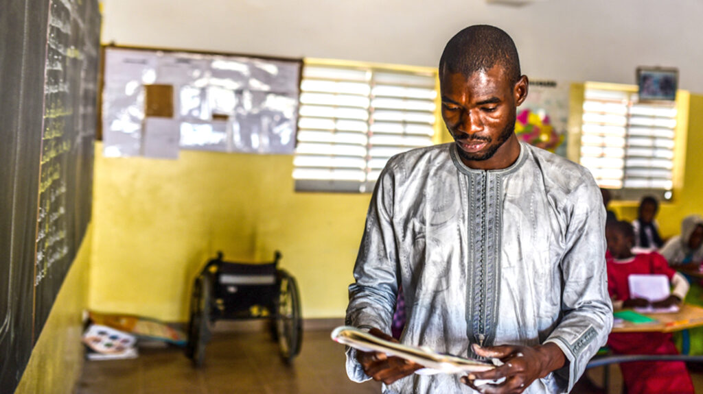 Teacher Soule looks at a workbook while standing at the blackboard. There's a wheelchair in the background, in the corner of the classroom.