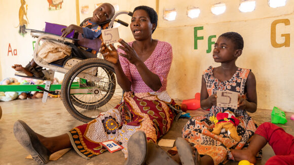 Nora teaches numbers to pre-school children. A child using a wheelchair sits on her left.