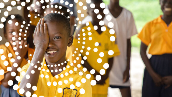 A school student wearing a bright yellow shirt smiles as he holds his right hand over his eye. There's an illustrated ring of small dots surrounding his face.