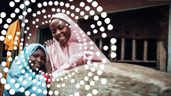 Asmau and Fatima, both wearing coloured headscarves, smile and embrace at their home in Nigeria.