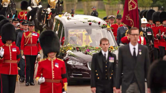 Queen Elizabeth's coffin is driven in a black hearse covered in flowers, flanked by Queens Guards wearing bearskins and full red military uniforms, plus officials in dark suits.