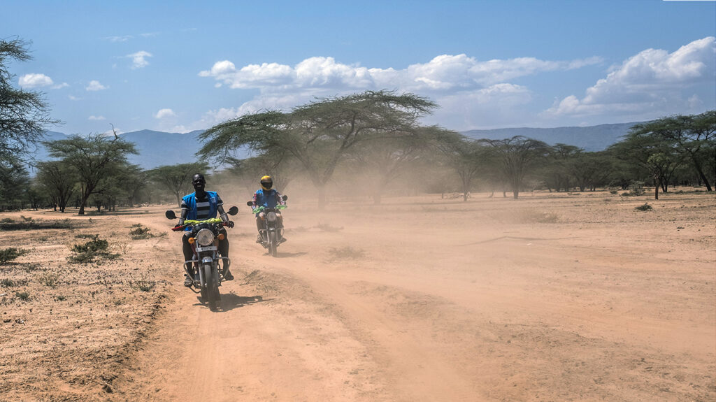 Two men ride motorbikes on a dusty dirt road, amid a spectacular rural landscape with distant trees and mountains.