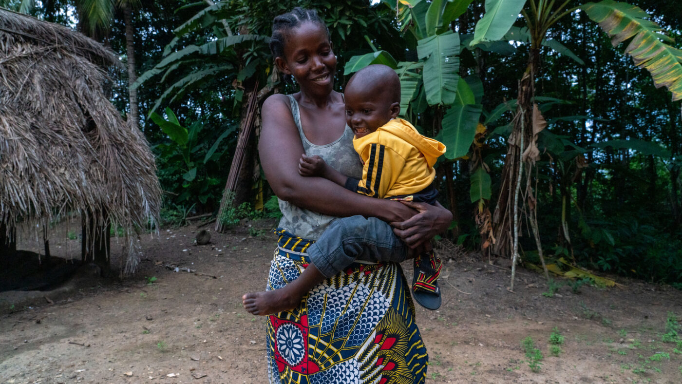 Mehnu's mother stands outside, holding Mehnu up. They are both smiling