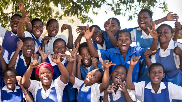 A large group of school students in Malawi. They're wearing bright blue uniforms and are gathered outside, smiling and cheering.