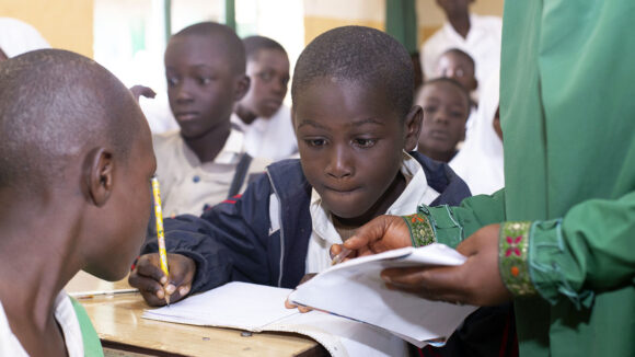 A young boy sits at a desk in a classroom, writing in an exercise book. A teacher stands next to him offering guidance.