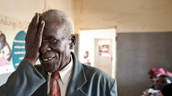 An older man smiles as he holds his right hand over his eye during an eye test.