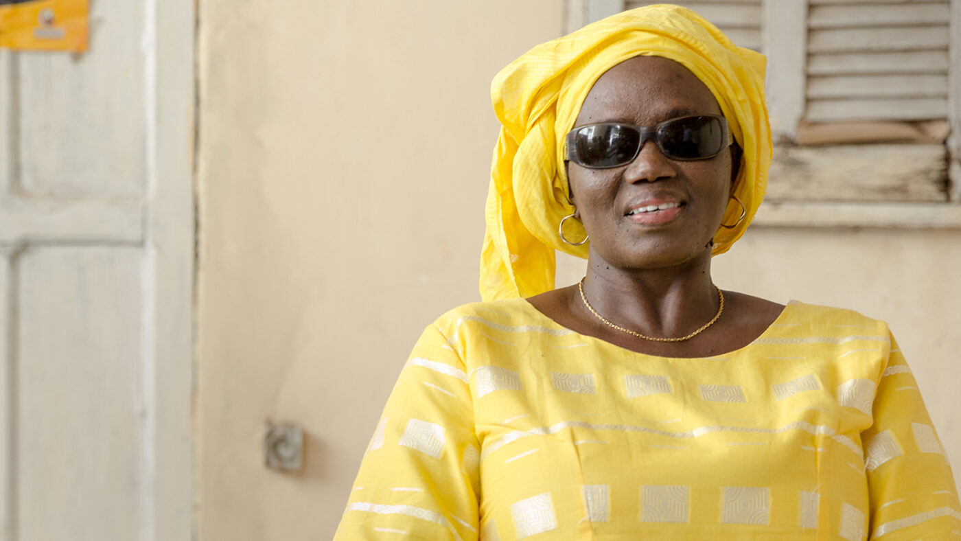 Seynabou, wearing dark glasses and a colourful yellow dress and headscarf.