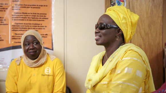 Seynabou sits with a woman who is wearing a yellow T-shirt with a Sightsavers logo.