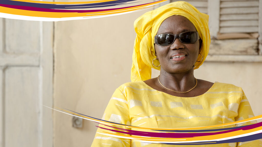 Seynabou, wearing dark glasses and a colourful yellow dress and headscarf.