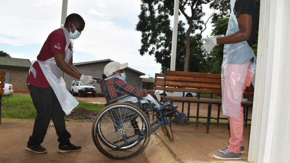 A man in a wheelchair is assisted by a health worker. There is no ramp to gain access into the building.