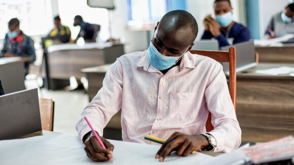 Daniel sits at a desk wearing a face mask and writing notes.