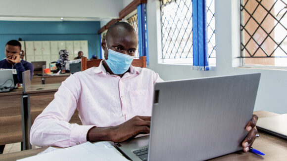 Daniel sits at a desk in a classroom. He is wearing a face mask and working on a laptop.