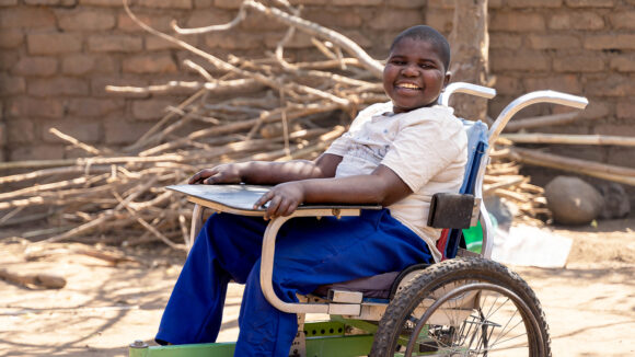 Enock sits in his wheelchair outside his home. He's wearing his school uniform and smiling broadly.