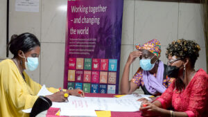 Three women from Cameroon sit around a table during a working group to support people with disabilities on gender-based violence. Behind them is a banner with information about the SDGs.