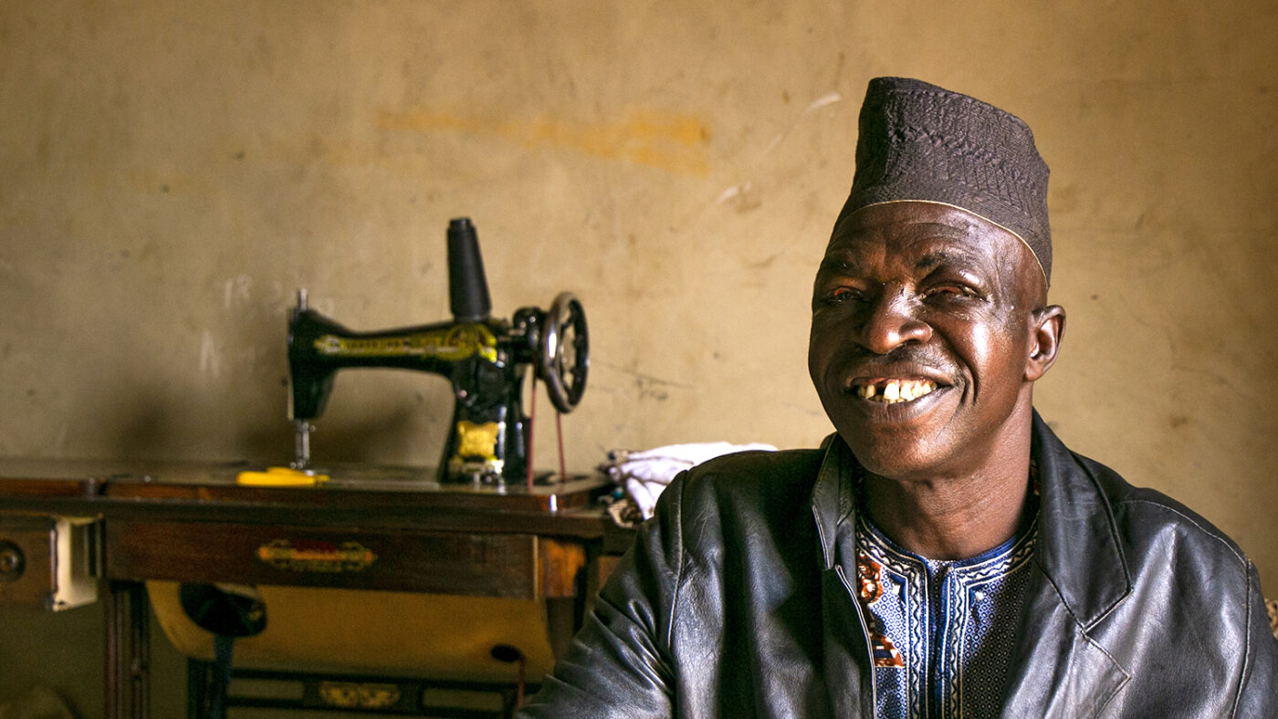 Fousseni smiles broadly. He's wearing a traditional African hat and grey tunic.