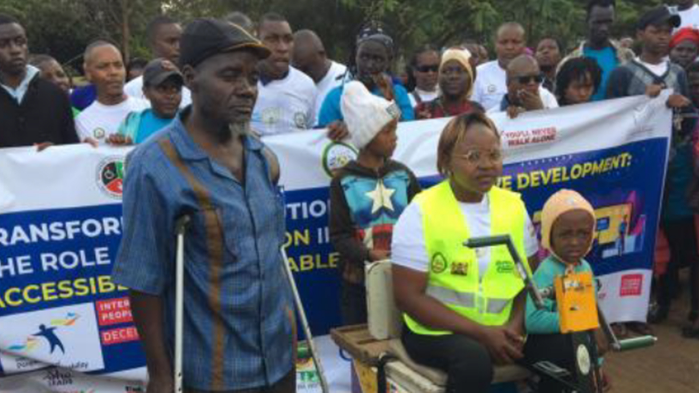 A man with crutches and a woman using a mobility scooter stand in front of a banner during International Day of People with Disabilities.
