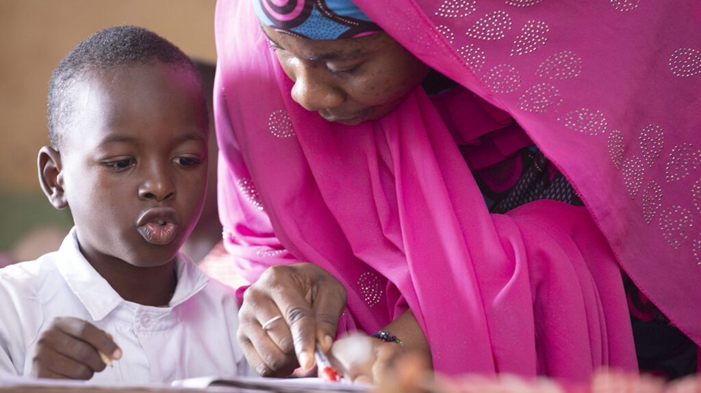 A teacher in Nigeria speaks to a child in the classroom. She's wearing a vibrant pink headscarf.
