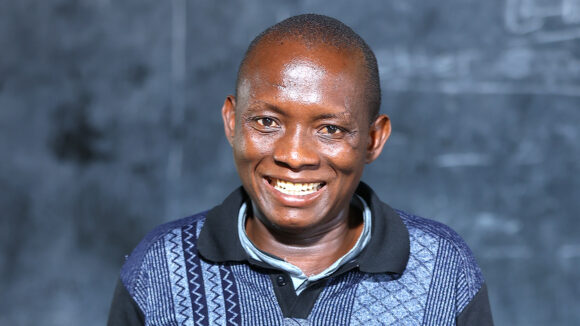 Teacher Bradon smiles at the camera with a blackboard in the background.
