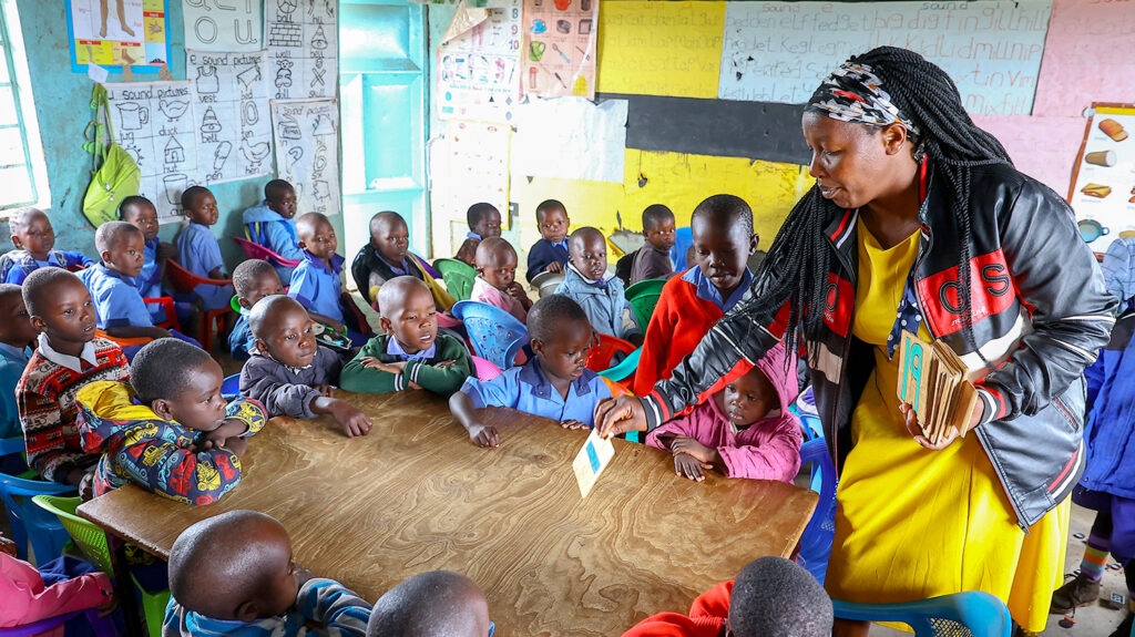 A classroom in Kenya, where a teacher hands out cards to students seated around a table.