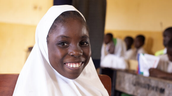 Hajara from Nigeria smiles broadly in her classroom. She's wearing a crisp white headscarf.