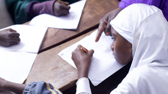 A student wearing a white headscarf writes on a piece of paper in the classroom.