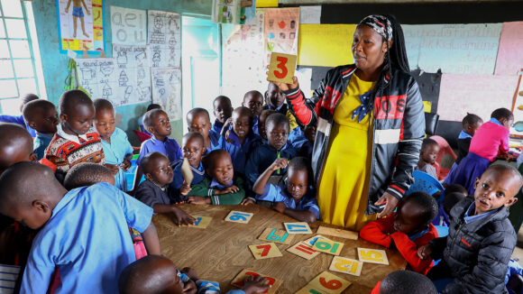 A classroom in Kenya, where a teacher shows a flashcard with the number three on it to students seated around a table.