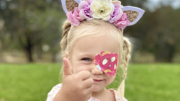 A young girl wearing a colourful eye patch smiles and holds her thumb up to the camera.