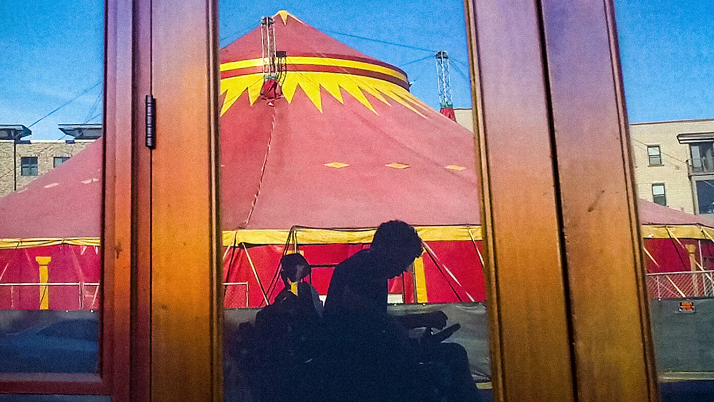 A still image from the film I Didn't See You There, showing the silhouette of a man in a wheelchair reflected in a doorway, with a colourful circus tent in the background.