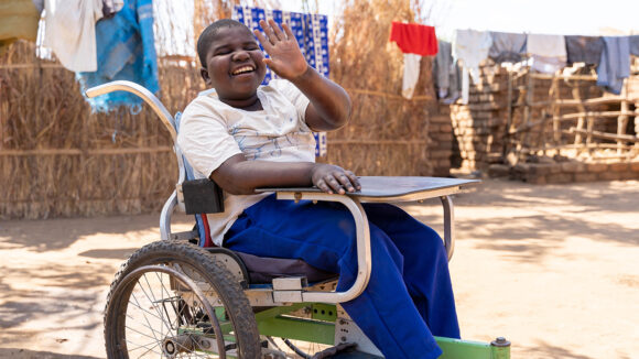 Enoch, who uses a wheelchair, smiles and waves at the camera.
