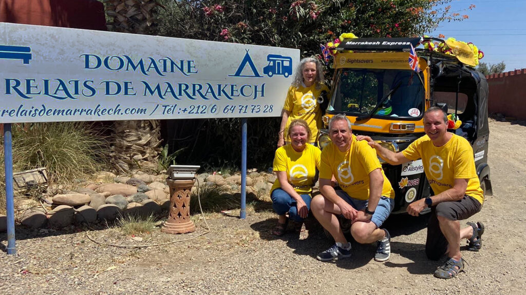 Paul and his team gather next to their yellow Sightsavers-branded tuk-tuk, wearing yellow Sightsavers t-shirts. They're standing next to a sign saying 'Domaine Relais de Marrakech'.