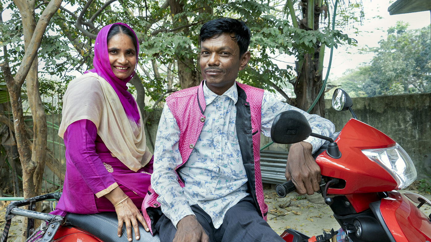 A husband and wife from Bangladesh sit on a motorbike, smiling at the camera.