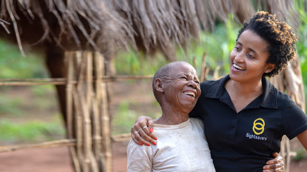 Mercia, wearing a black Sightsavers t-shirt, hugs patient Maria outside in a rural Mozambique village. They're both smiling.