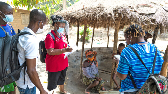 A group of researchers, carrying backpacks and holding paperwork, speak to local villagers outdoors in Sierra Leone.