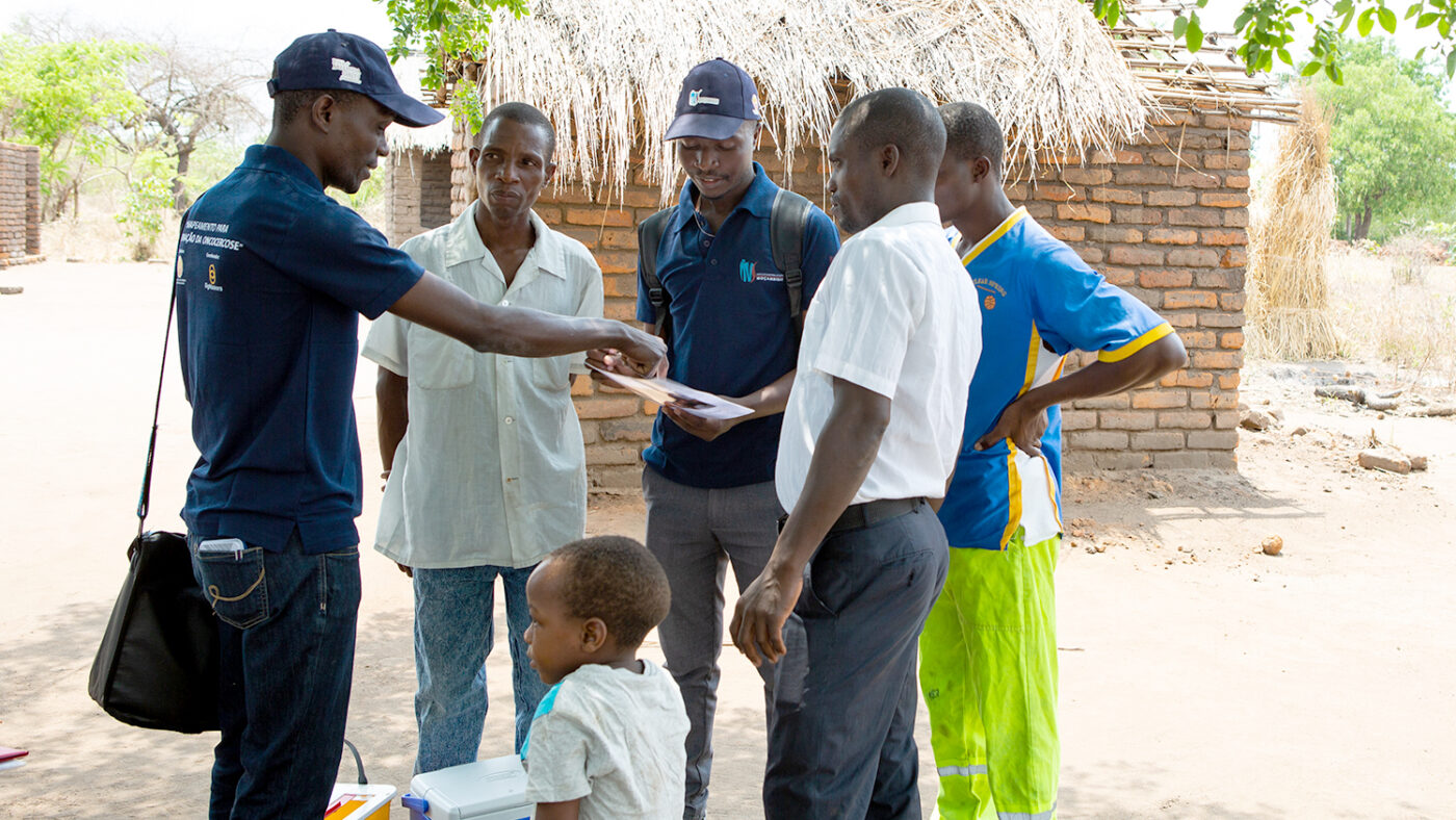 In a village in Mozambique, two people in blue polo shirts hold clipboards and talk to members of the local community.