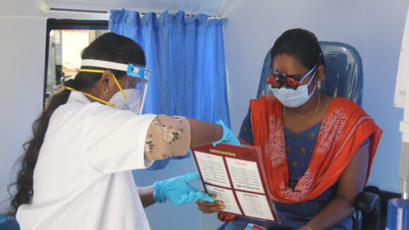 An eye health worker in a clinic holds up an eye test chart for a patient who is having her eyes screened.