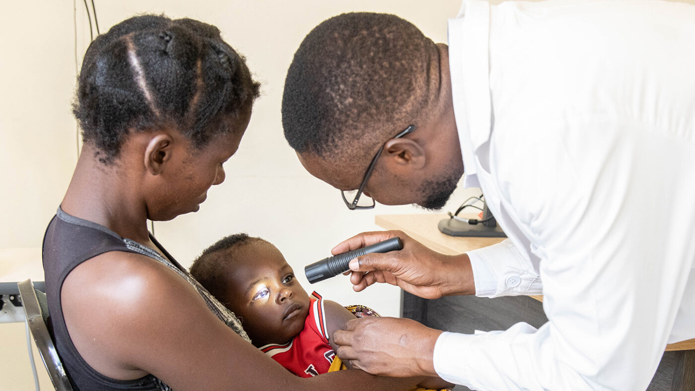 An eye health worker wearing white coat shines a light into a young child's eyes as he sits on his mother's lap.