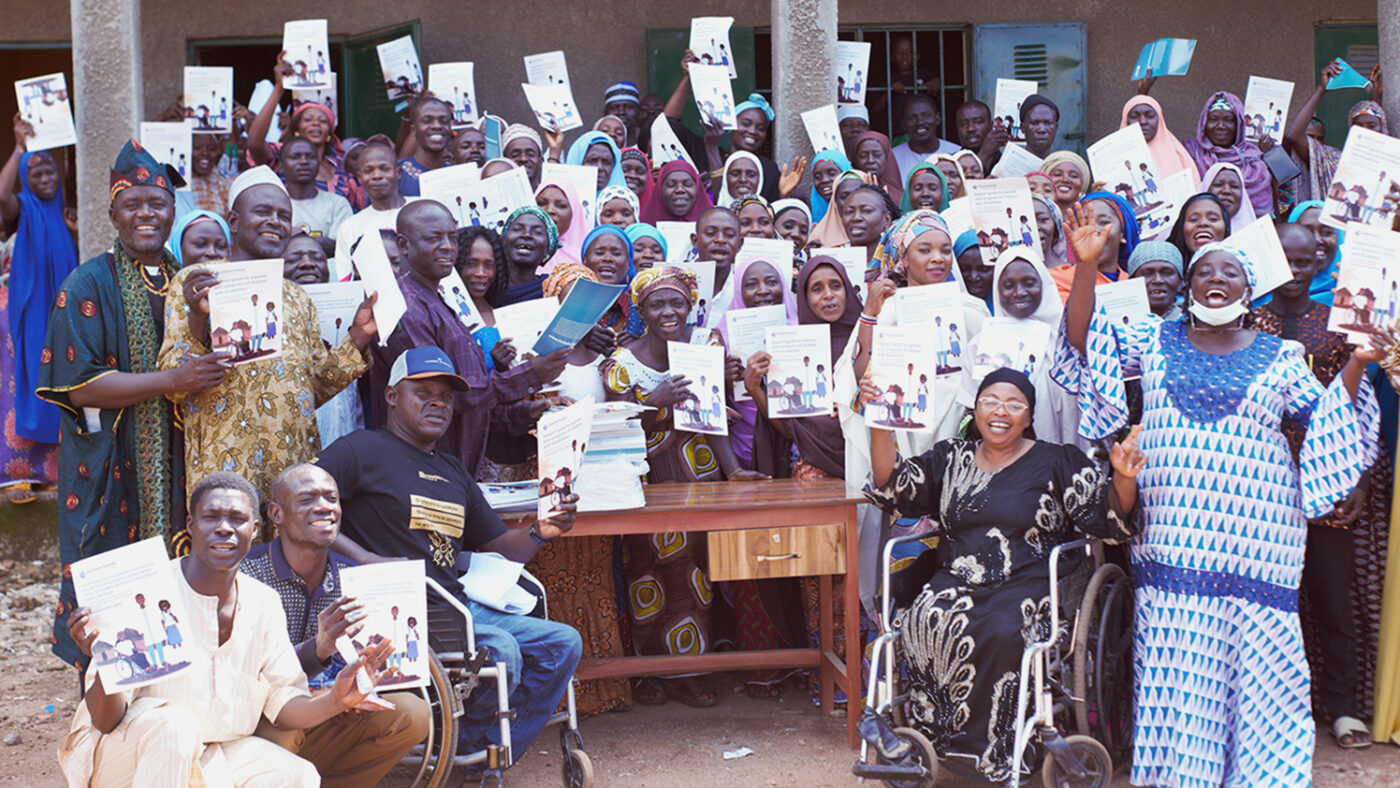 A large group of people in Nigeria hold up copies of a guide for parents of children with disabilities information about how to support their children’s education.