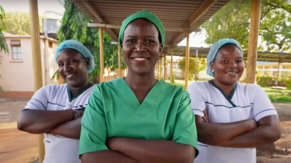 Three eye health staff wearing surgical scrubs and hats outside a clinic in Uganda.