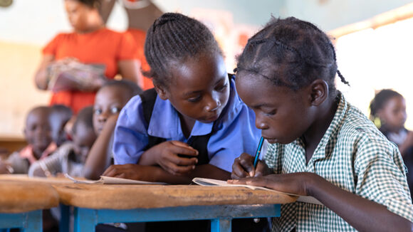 Two school students in Zambia huddle together at a desk. One is writing, the other is looking at the work.
