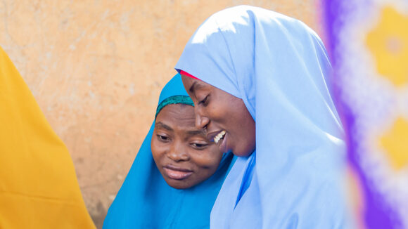 Two women wearing colourful headscarves lean together and smile.