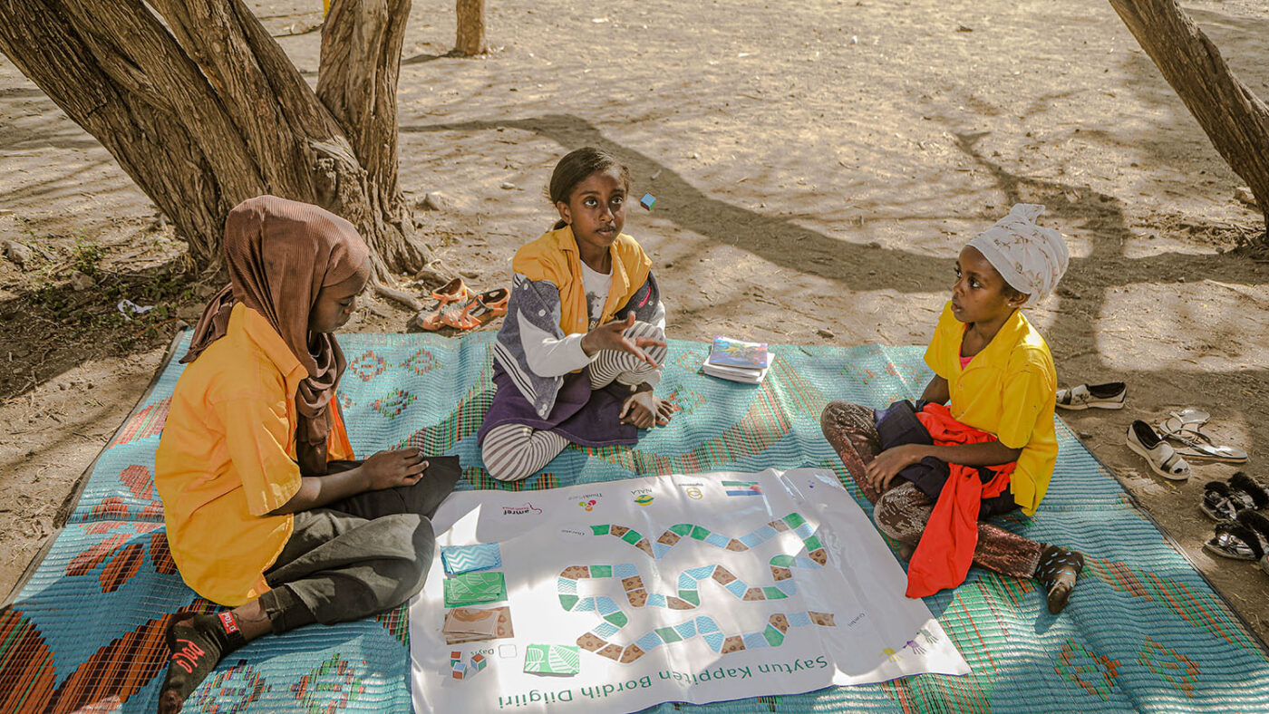 Three girls sit and play a board game laid out on the ground.