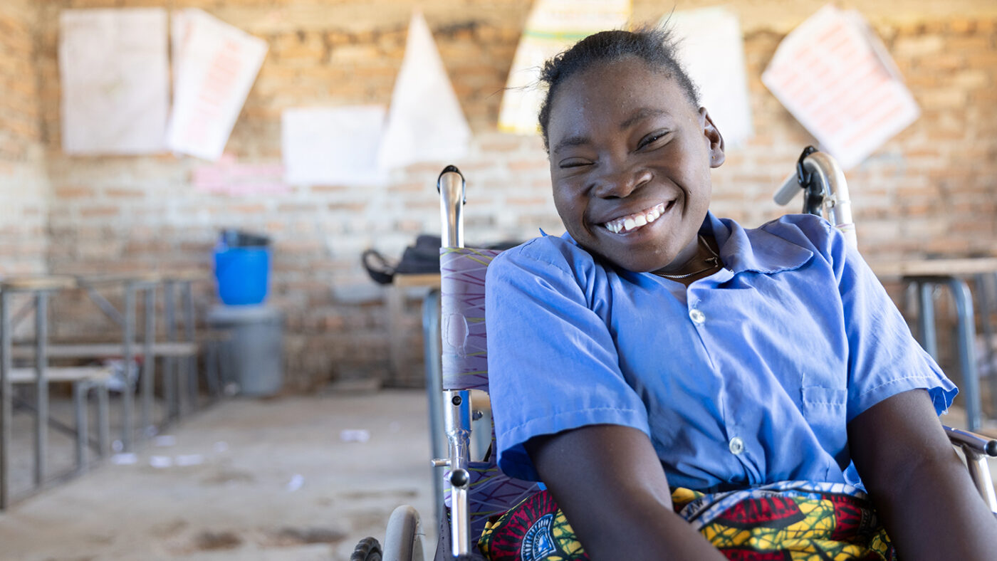 A girl smiles in a classroom in Zambia. She's sitting in a wheelchair and wearing a blue school shirt.