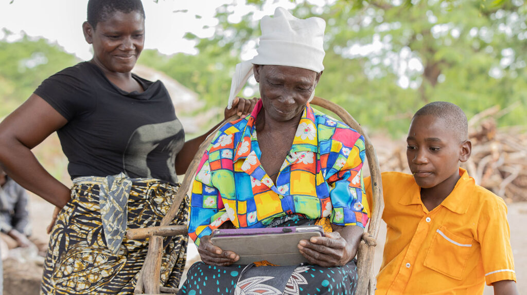 Angeshita watches a video of herself on a tablet. Her daughter and grandson sit beside her.