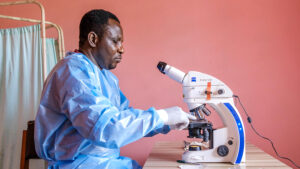 A laboratory scientist wearing surgical gear sits at a table using a microscope.